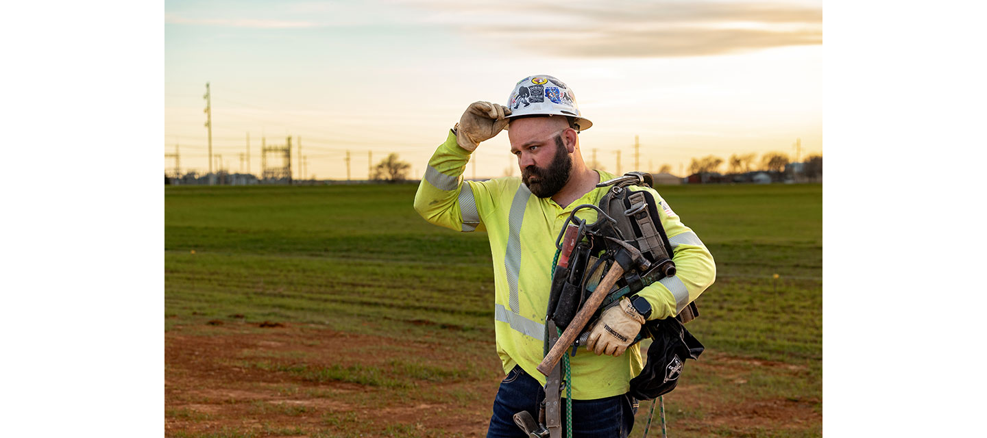 Un liniero, Kevin Walker, con casco y camisa de seguridad amarilla, se seca la frente mientras sostiene un cinturón de herramientas sobre su hombro en un campo de hierba al atardecer.